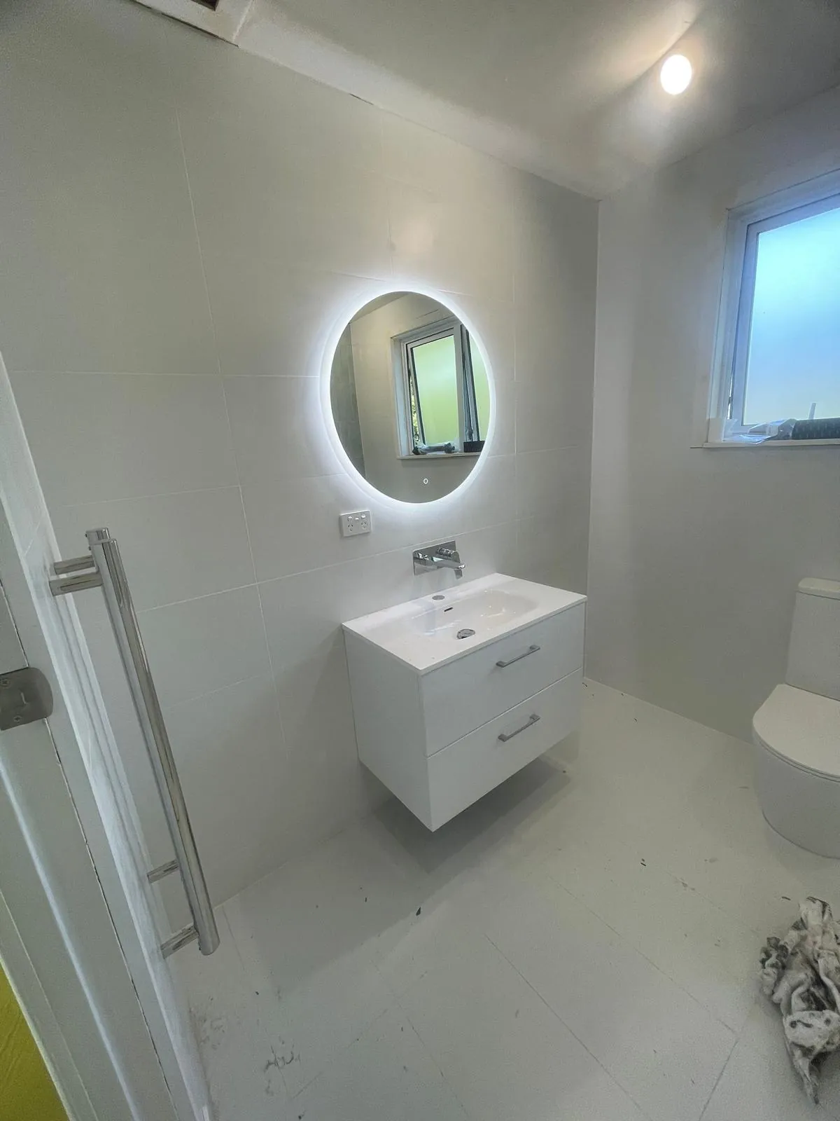A finished bathroom with a backlit round mirror, wall-hung vanity and a ceiling downlight
