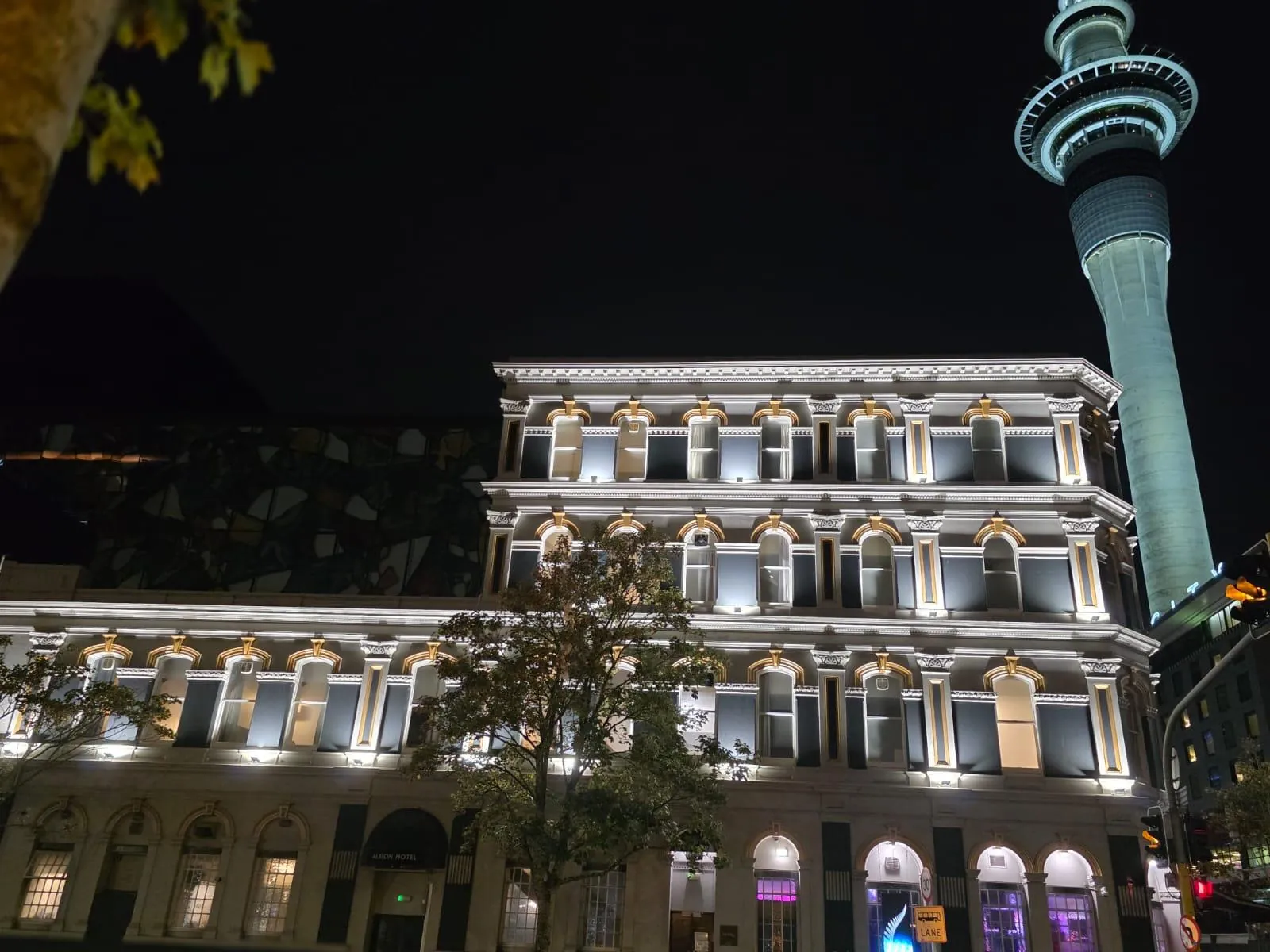 A heritage Auckland building lit at night by architectural facade lighting, with the Sky Tower behind